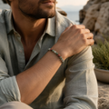 Mann präsentiert das African Bloodstone Natursteinarmband mit 6 mm großen grünen, grauen und roten Natursteinperlen in einer sonnigen, felsigen Küstenlandschaft.