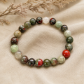 African Bloodstone Natursteinarmband mit 8 mm großen, individuell gemusterten Perlen in Dunkelgrün, Grau, Rot und Creme auf hellem Leinen mit getrockneten Gräsern präsentiert.