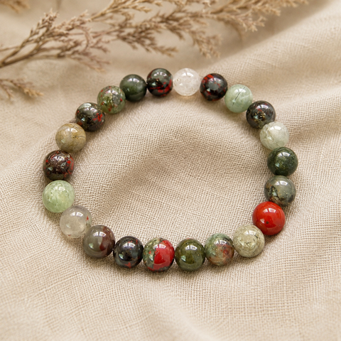 African Bloodstone Natursteinarmband mit 8 mm großen, individuell gemusterten Perlen in Dunkelgrün, Grau, Rot und Creme auf hellem Leinen mit getrockneten Gräsern präsentiert.