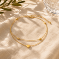 Elegante goldfarbene Fußkette mit doppelter Kettenführung, feinen Kugeldetails und Herzanhänger. Stilvoll präsentiert auf hellem Leinenstoff mit warmem Sonnenlicht, sanften Schatten und mediterraner Sommeratmosphäre. Femininer Schmuck für Alltag, Urlaub und besondere Anlässe.