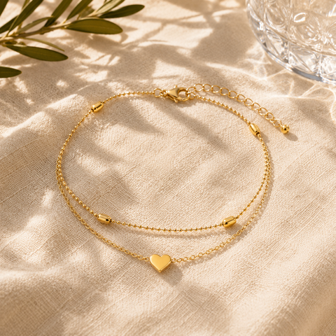 Elegante goldfarbene Fußkette mit doppelter Kettenführung, feinen Kugeldetails und Herzanhänger. Stilvoll präsentiert auf hellem Leinenstoff mit warmem Sonnenlicht, sanften Schatten und mediterraner Sommeratmosphäre. Femininer Schmuck für Alltag, Urlaub und besondere Anlässe.