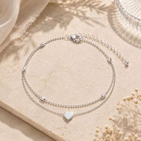 Filigrane Amora Fußkette in Silber mit doppelter Kettenführung, Herzanhänger und feinen Kugeldetails. Stilvoll inszeniert auf hellem Naturstein mit weichem Leinenstoff, zarten Blüten, warmem Sonnenlicht und mediterraner Sommeratmosphäre. Zeitloser Schmuck für Alltag, Urlaub und besondere Anlässe.
