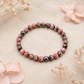 Filigranes Black Veined Rhodonite Natursteinarmband mit 6 mm großen Perlen in Rosa, Altrosa, Braun und Schwarz auf hellem Leinen mit dezenten Naturblumen.