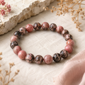 Black Veined Rhodonite Natursteinarmband mit 8 mm großen, individuell gemusterten Perlen in Rosa-, Braun- und Schwarztönen auf hellem Naturstoff mit floralen Akzenten.
