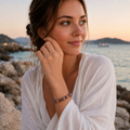 Frau trägt das Black Veined Rhodonite Natursteinarmband mit 8 mm großen rosa-schwarz gemusterten Perlen am Handgelenk in einer warmen Küstenkulisse bei Sonnenuntergang.