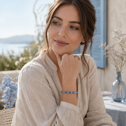 Frau trägt das Blue Aventurine Natursteinarmband mit 6 mm großen, etwas dunkleren blauen und grauen Perlen am Handgelenk in einer hellen mediterranen Kulisse am Wasser.