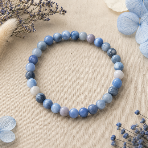 Filigranes Blue Aventurine Natursteinarmband mit 6 mm großen Perlen in Hellblau, Mittelblau, Grau und zartem Lavendel auf hellem Leinen mit dezenten blauen Trockenblumen.