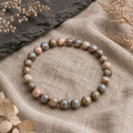 Blue Que Sera Opal Natursteinarmband mit 6 mm großen, individuell gemusterten Perlen in sanftem Blau, Rosa, Beige und Grau auf hellem Naturstoff mit dunklem Stein und Trockenblumen präsentiert.