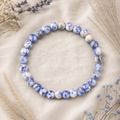 Filigranes Blue Spot Jasper Natursteinarmband mit 6 mm großen, blau-weiß gemusterten Perlen auf hellem Leinen mit dezenten Trockenblumen in passenden Farbtönen.
