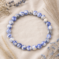Blue Spot Jasper Natursteinarmband mit 8 mm großen, individuell marmorierten Perlen in Blau, Creme und Grau auf hellem Naturstoff mit farblich abgestimmten Gräsern und Blumen.