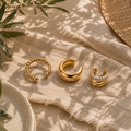 Vergoldetes Ear Cuff Set Camille mit Kristallbesatz bestehend aus drei modernen Ear Cuffs in Gold. Eleganter Ohrschmuck auf beigem Stoff mit mediterranem Sommer-Ambiente und natürlichen Grüntönen.