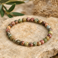 Filigranes Cherry Greek Jasper Armband mit 6 mm großen Natursteinperlen in Grün-, Rot-, Braun- und Sandtönen, hochwertig auf einem natürlichen Stein mit dezenten Pflanzen und warmem Licht inszeniert.