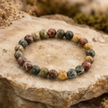 Cherry Greek Jasper Armband mit 8 mm großen, polierten Natursteinperlen in erdigen Grün-, Rot-, Braun- und Beigetönen, natürlich auf einer rauen Steinoberfläche mit Pflanzen präsentiert.