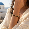 Frau präsentiert das Cherry Greek Jasper Armband mit 8 mm großen Natursteinperlen am Handgelenk vor einer hellen griechischen Küstenkulisse mit weißer Architektur und blauem Meer.