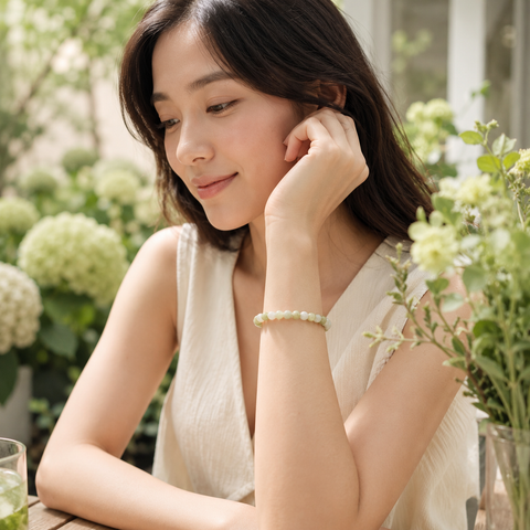 Moderne asiatische Frau trägt das Flower Jade Natursteinarmband mit 6 mm großen weiß-grünen Perlen am Handgelenk in einer hellen Gartenumgebung mit grünen und cremefarbenen Blüten.