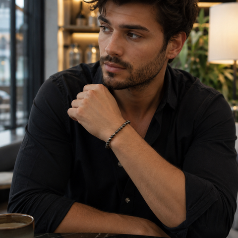 Mann mit schwarzem Hemd trägt das filigrane Gold Obsidian Natursteinarmband mit 6 mm großen dunklen Perlen in einem modernen, warm beleuchteten Café.