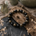 Gold Obsidian Natursteinarmband mit 8 mm großen, tiefschwarzen und bronzefarben schimmernden Perlen auf dunklem Stein, Holz und dezenten Naturelementen präsentiert.
