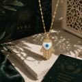 Goldfarbene Hand der Fatima Halskette mit blauem Nazar-Auge und funkelnden Details auf hellem Steinpodest. Luxuriöse Schmuckinszenierung mit grünen Stoffelementen, orientalischen Mustern und warmem Licht im AVRE Studios Stil.
