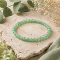 Green Aventurine Natursteinarmband mit 6 mm großen, leicht transparenten Perlen in sanftem Grün, natürlich auf hellem Stein mit Eukalyptus, cremefarbenen Blüten und grünem Stoff präsentiert.