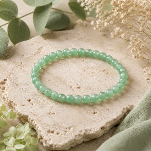 Green Aventurine Natursteinarmband mit 6 mm großen, leicht transparenten Perlen in sanftem Grün, natürlich auf hellem Stein mit Eukalyptus, cremefarbenen Blüten und grünem Stoff präsentiert.