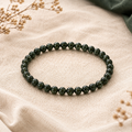 Filigranes Green Sandstone Natursteinarmband mit 6 mm großen, dunkelgrünen und leicht schimmernden Perlen auf hellem Leinen mit dezenten Trockenblumen und dunkelgrünem Stoffdetail.