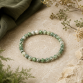 Green Spot Jasper Natursteinarmband mit 6 mm großen, grün-weiß gemusterten Perlen auf einer hellen beigefarbenen Oberfläche mit dunkelgrünem Stoff und dezenten Pflanzenelementen.