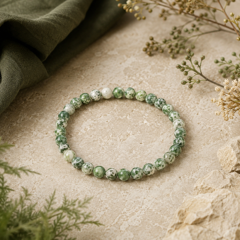 Green Spot Jasper Natursteinarmband mit 6 mm großen, grün-weiß gemusterten Perlen auf einer hellen beigefarbenen Oberfläche mit dunkelgrünem Stoff und dezenten Pflanzenelementen.