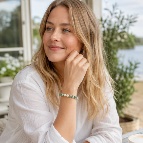 Blonde Frau trägt das Green Spot Jasper Natursteinarmband mit 8 mm großen grün-weißen Perlen zu einer weißen Bluse in einem hellen skandinavischen Gartencafé am Wasser.