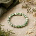 Green Spot Jasper Natursteinarmband mit 8 mm großen, individuell gemusterten Perlen in Grün, Creme und Weiß, natürlich auf hellem Beige mit dunkelgrünem Stoff und botanischen Details präsentiert.