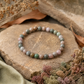 Indian Agate Natursteinarmband mit 6 mm großen, individuell gemusterten Perlen in gedecktem Grün, Rosa, Beige, Grau und Braun auf einer beigefarbenen Naturfläche mit farblich abgestimmten Trockenpflanzen.