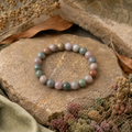 Indian Agate Natursteinarmband mit 8 mm großen, polierten Perlen in sanften Grün-, Rosa-, Grau-, Beige- und Brauntönen, natürlich auf strukturiertem Beige mit dezenten botanischen Elementen inszeniert.