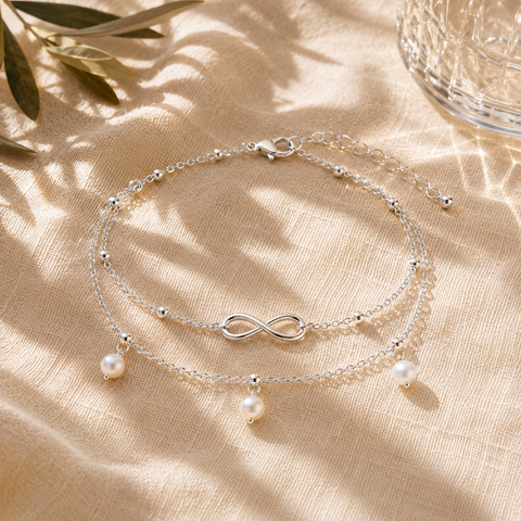 Elegante silberfarbene Infinity-Fußkette mit doppelter Kette, drei weißen Perlenanhängern und filigranen Silberkugeln. Stilvoll inszeniert auf cremefarbenem Leinenstoff mit mediterranen Lichtreflexionen und sommerlicher Premium-Atmosphäre. Ideal für Alltag, Urlaub und besondere Anlässe.