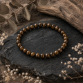 Iron Tiger Eye Natursteinarmband mit 6 mm großen, polierten Perlen in Braun, Goldbraun und dunklen Erdtönen auf schwarzem Naturstein mit Holz und Trockenblumen präsentiert.