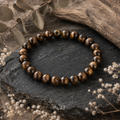 Iron Tiger Eye Natursteinarmband mit 8 mm großen Perlen und charakteristischer braun-goldener Maserung auf dunklem Stein zwischen verwittertem Holz und dezenten Naturelementen.