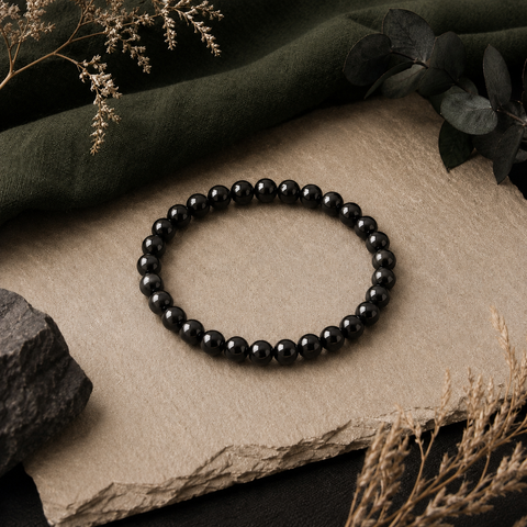 Java Black Obsidian Armband mit 6 mm großen, tiefschwarzen und glänzend polierten Perlen auf einer beigefarbenen Steinfläche mit dunkelgrünem Stoff und getrockneten Pflanzen.