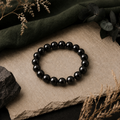 Java Black Obsidian Armband mit 8 mm großen, gleichmäßig schwarzen und polierten Natursteinperlen auf beigem Stein, ergänzt durch dunkelgrünen Stoff und natürliche Trockenblumen.