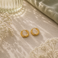Goldfarbene Luna Creolen in 12 mm Größe auf hellem Naturstein mit sanften Lichtreflexen, Glasvase, salbeigrünem Stoff und feinen weißen Blüten im eleganten AVRE Studios Stil.