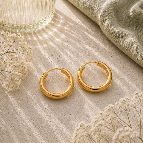Goldfarbene Luna Creolen in 18 mm Größe auf hellem Naturstein mit sanften Lichtreflexen, eleganter Glasvase, salbeigrünem Stoff und feinen weißen Blüten im hochwertigen AVRE Studios Stil.