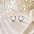 Luna Creolen in Silber mit 10 mm Durchmesser aus hochwertigem Edelstahl. Minimalistische Creolen mit glänzender Oberfläche, stilvoll präsentiert auf hellem Stoff mit Glasreflexionen und feinen weißen Blüten im Hintergrund. Ideal für einen zeitlosen und eleganten Look.