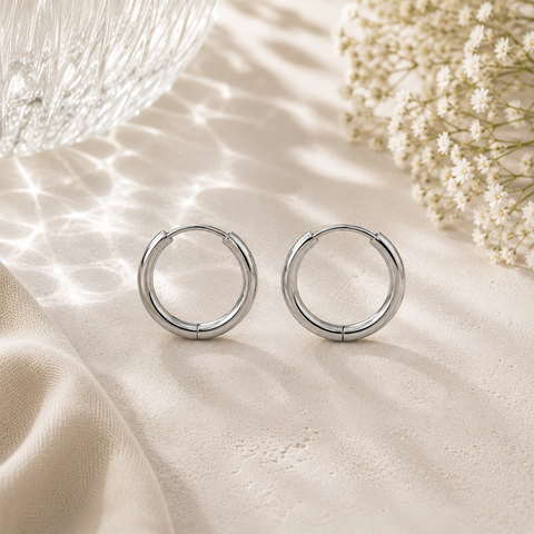 Luna Creolen in Silber mit 14 mm Durchmesser aus hochwertigem Edelstahl. Klassische mittelgroße Creolen mit glänzender Oberfläche, stilvoll inszeniert auf heller Natursteinoptik mit Glasreflexionen, feinen weißen Blüten und elegantem Stoff im Hintergrund.