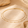 Elegante goldfarbene Mira Bauchkette von AVRE Studios mit doppelreihigem Design und feinen Kugeldetails auf einer warmen Naturstein-Oberfläche präsentiert. Moderne Taillenkette für stilvolle Sommer- und Urlaubslooks.