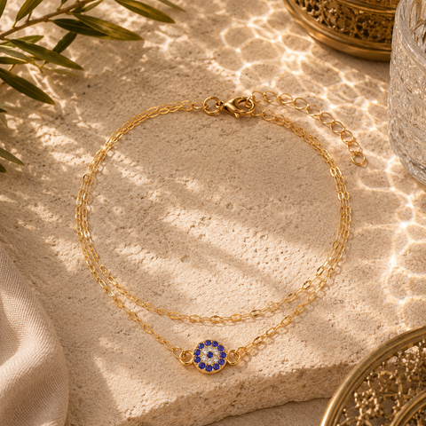 Elegante Safira Fußkette in Gold mit funkelndem Nazar Auge als traditionellem Schutzsymbol und Glücksbringer. Hochwertig inszeniert auf hellem Naturstein mit orientalischen Ornamenten, mediterranen Lichtreflexionen und warmem Sonnenlicht. Filigraner Sommerschmuck für stilvolle Alltagslooks, Urlaub und besondere Anlässe.