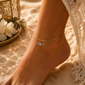 Frau trägt die Nayla Fußkette in Gold mit blauem Nazar Auge am Knöchel. Die feine doppelte Gliederkette verleiht dem Schmuckstück eine elegante Ausstrahlung und wird in einer warmen Sommerkulisse mit goldenem Licht, Sand und orientalischen Akzenten stilvoll in Szene gesetzt. Ideal für stilvolle Alltagslooks und besondere Momente.