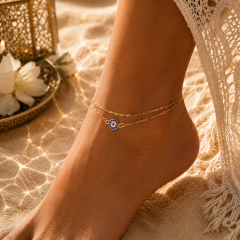 Frau trägt die Nayla Fußkette in Gold mit blauem Nazar Auge am Knöchel. Die feine doppelte Gliederkette verleiht dem Schmuckstück eine elegante Ausstrahlung und wird in einer warmen Sommerkulisse mit goldenem Licht, Sand und orientalischen Akzenten stilvoll in Szene gesetzt. Ideal für stilvolle Alltagslooks und besondere Momente.
