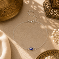 Filigrane silberfarbene Nazar Fußkette mit blauem Auge als orientalischem Schutzsymbol. Stilvoll inszeniert auf hellem Naturstein mit traditionellen Ornamenten, feinem Stoff, zarten Blüten und warmem Sonnenlicht. Eleganter Sommerschmuck mit mediterran-orientalischer Ausstrahlung für Alltag und besondere Anlässe.