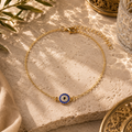 Filigrane goldfarbene Nazar Luxe Fußkette mit funkelndem blauem Schutzauge und feiner Gliederkette. Hochwertig inszeniert auf hellem Naturstein mit mediterranen Lichtreflexionen, orientalischen Akzenten und warmer Sommeratmosphäre. Eleganter Schmuck für Alltag, Urlaub und besondere Anlässe.