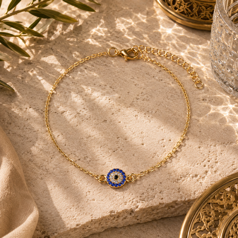 Filigrane goldfarbene Nazar Luxe Fußkette mit funkelndem blauem Schutzauge und feiner Gliederkette. Hochwertig inszeniert auf hellem Naturstein mit mediterranen Lichtreflexionen, orientalischen Akzenten und warmer Sommeratmosphäre. Eleganter Schmuck für Alltag, Urlaub und besondere Anlässe.