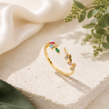 Goldring mit bunten funkelnden Kristallen auf hellem Naturstein. Umgeben von cremefarbenem Satinstoff und grünen Pflanzen in einer hochwertigen, warmen Schmuckszene.