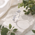 Filigraner offener Silberring mit funkelnden floralen Kristallen auf hellem Naturstein. Umgeben von cremefarbenem Satin und grünen Pflanzen in einer luxuriösen, natürlichen Schmuckinszenierung.
