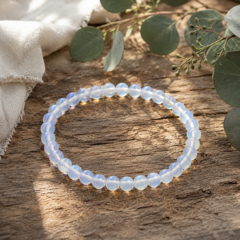 Opalite-Armband mit 6 mm großen, transparent-milchigen Perlen und sanftem blauem Schimmer auf rustikalem Holz, ergänzt durch helles Leinen und natürliche Eukalyptuszweige.